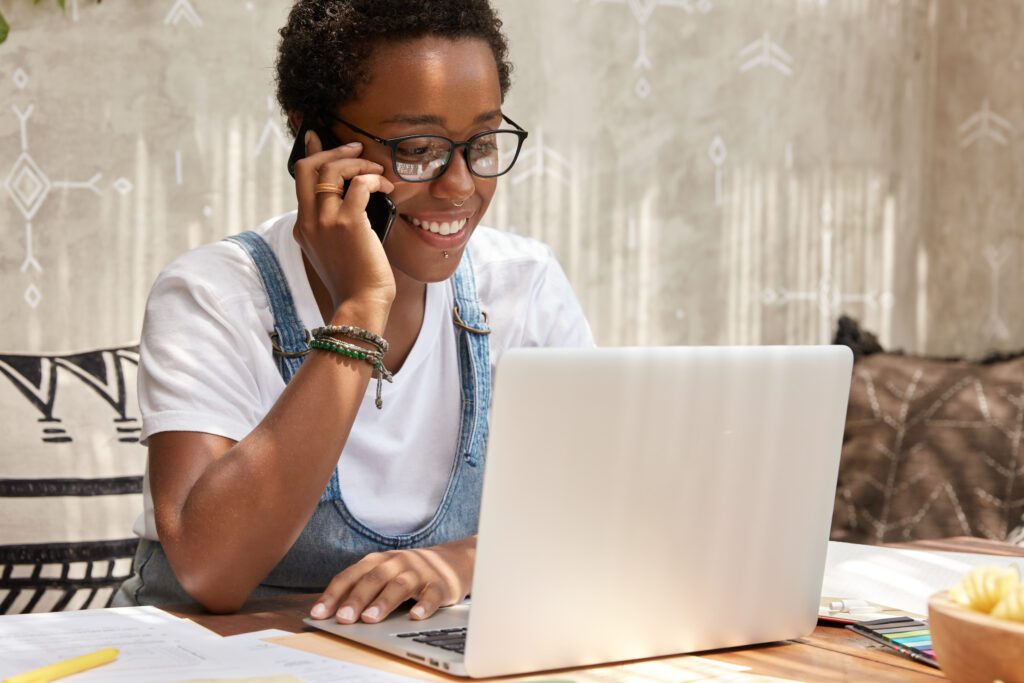 Stylish African American woman calls on smart phone looks in laptop computer, updates software, has