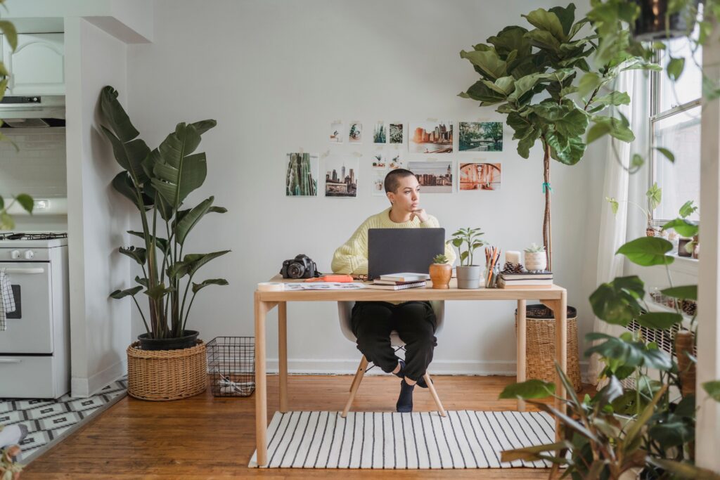 Full body of female freelancer looking at window while sitting at table and working on laptop in spacious room