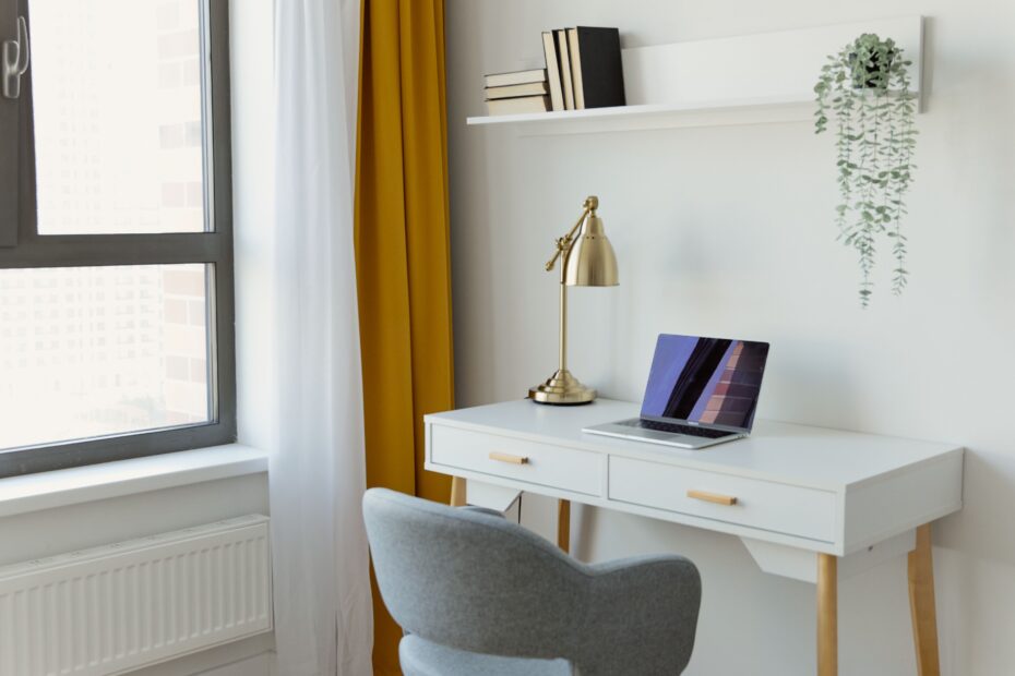 A modern home office with a laptop on a white desk, enhanced by natural light from a large window and minimalist decor.