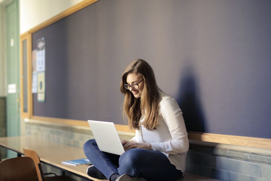A woman seated on a table in a classroom works on her laptop, embodying modern education.